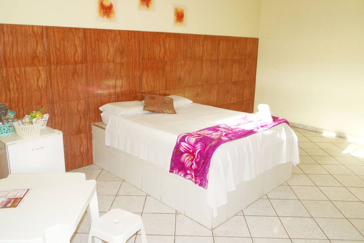 A hotel room with a wooden headboard, a bed with white linens and a pink floral blanket, a small white desk, and a chair.