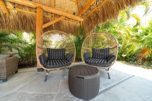 Two wicker chairs with black cushions under a thatched roof, a round wicker table, and a stone floor.