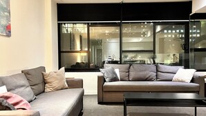 Una sala de estar moderna con un sofá gris, una mesa de centro y un amplio ventanal con vistas a un paisaje urbano.