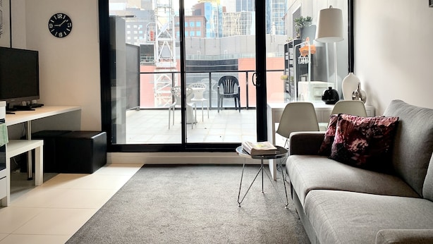 Una sala de estar moderna con un sofá gris, un televisor de pantalla plana, una mesita de centro pequeña y un amplio ventanal corredero que da a un balcón exterior con vistas a la ciudad.