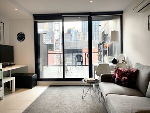 Un salon moderne avec un canapé gris, un téléviseur à écran plat, une petite table basse et une grande porte-fenêtre coulissante donnant sur un balcon extérieur avec vue sur la ville.