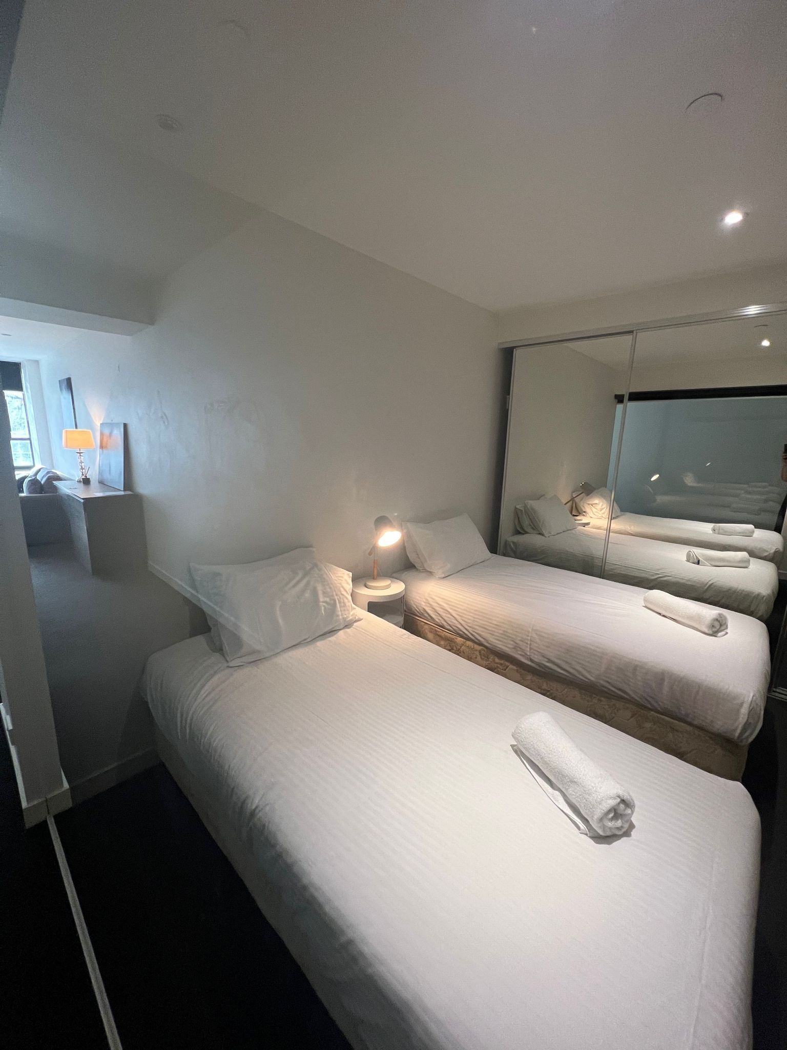 Habitación de hotel con dos camas, una mesita de noche con lámpara, un espejo grande y vistas al exterior.