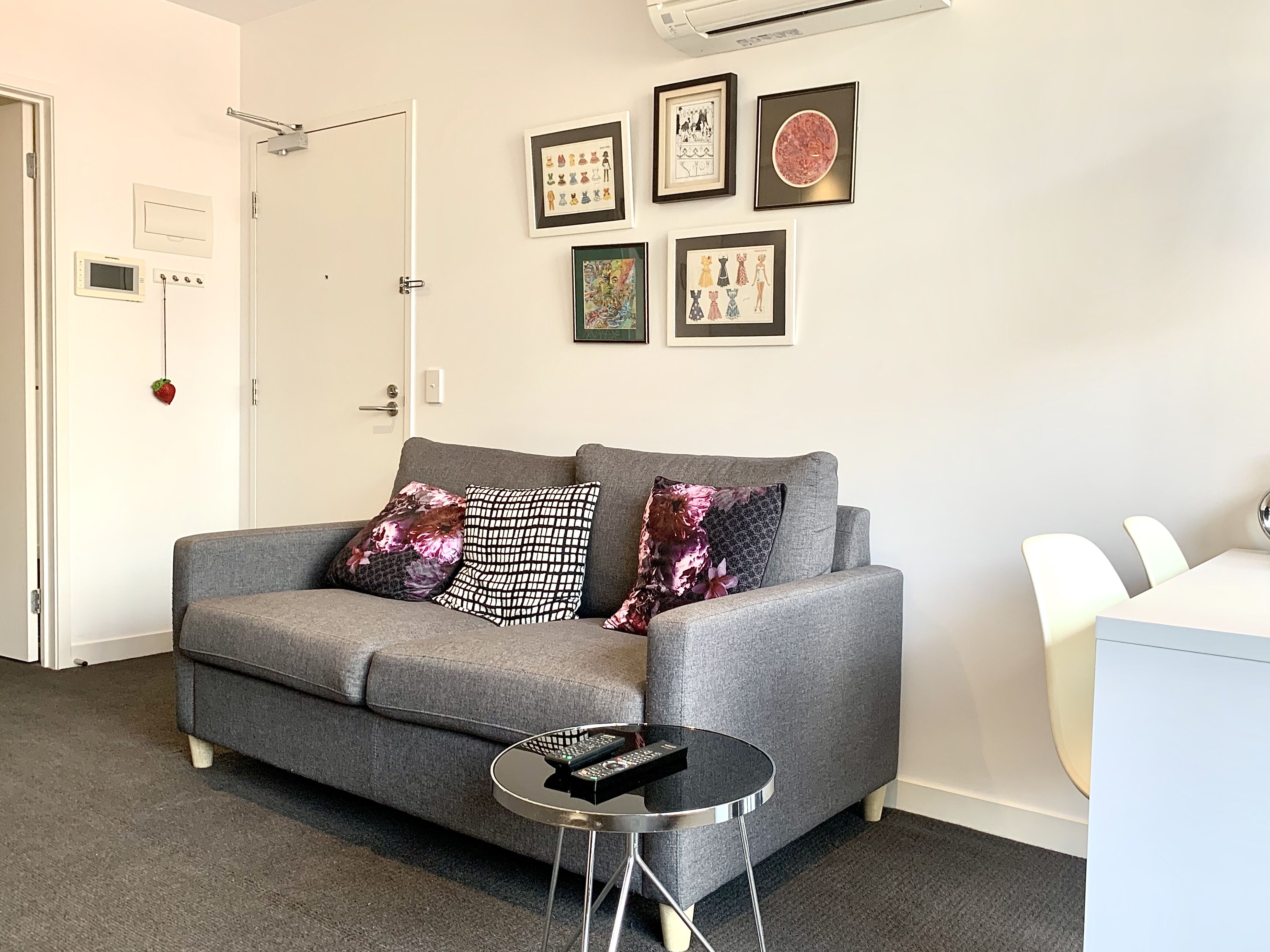 A grey sofa with patterned cushions, a small round table with a remote control, and framed artwork on the wall.