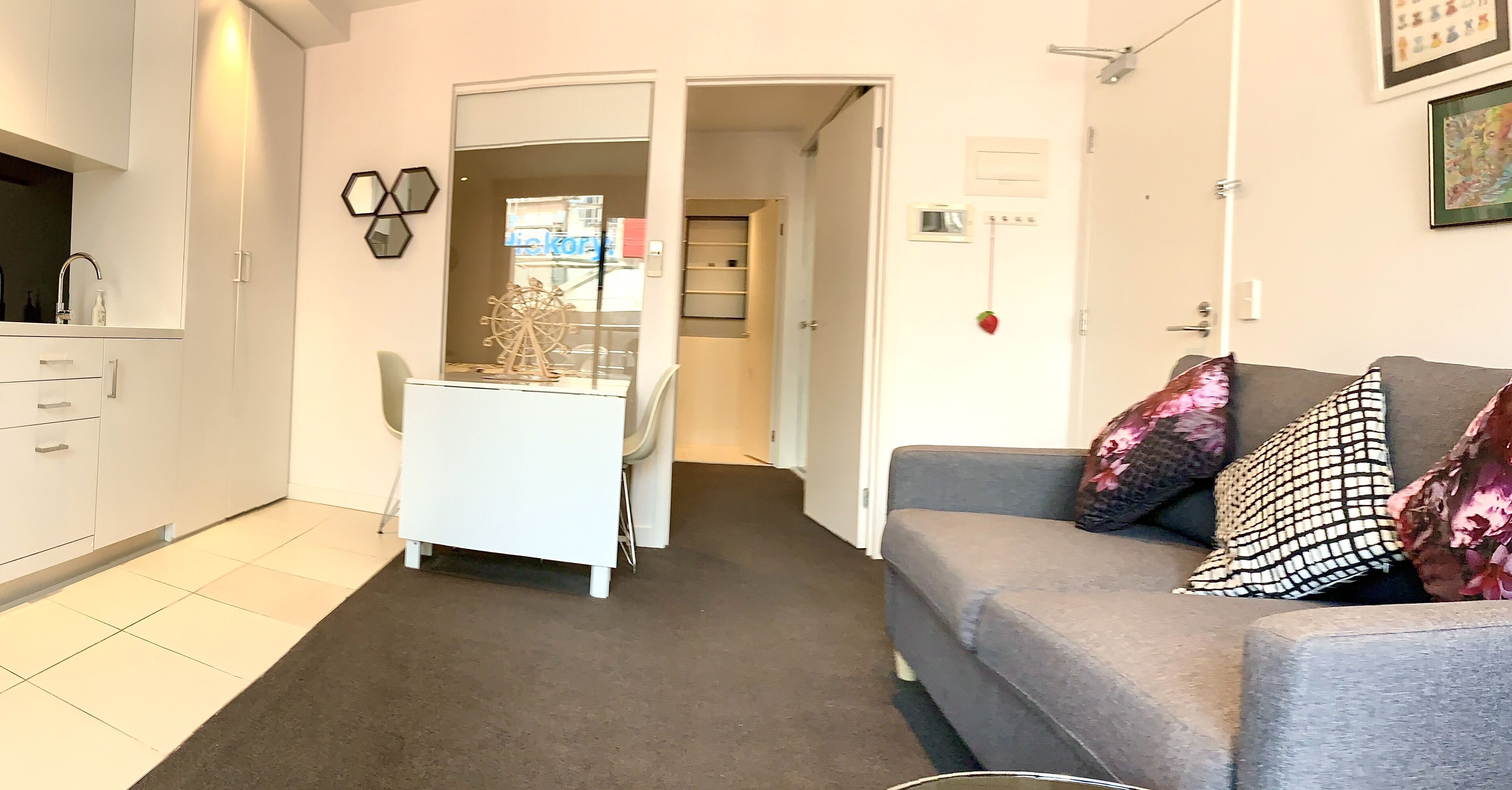 A compact living space with a kitchenette, a grey sofa with patterned cushions, and a white desk with a decorative wheel.