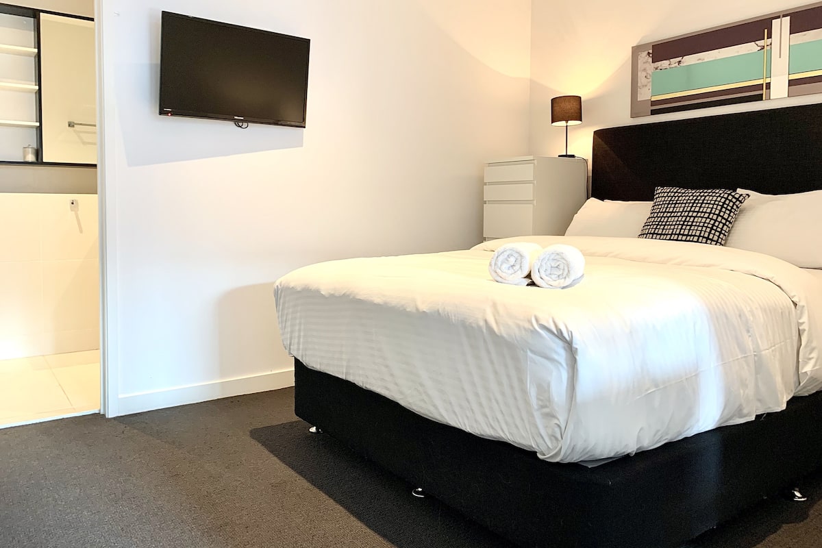 A modern bedroom with a large bed, a flat-screen TV mounted on the wall, a bedside lamp, and a window with a view of the sky.