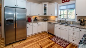 A modern kitchen with white cabinets, a stainless steel refrigerator, a coffee maker, and a window with a view of trees. - Beautiful New Home!! (Intervale)