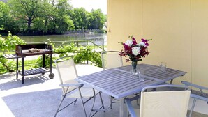 Une terrasse extérieure avec une table et des chaises, un petit chariot, et une vue sur une rivière et des arbres.