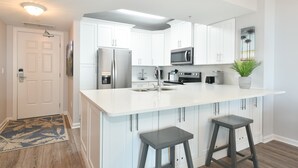 Una cocina moderna con armarios blancos, un refrigerador de acero inoxidable, un microondas y una cafetera. Hay dos taburetes de bar y una planta en maceta.