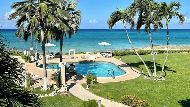 A pool area with lounge chairs and umbrellas, surrounded by palm trees and a view of the ocean. - Oceanfront 2 BR & Pool - Cocoplum #8 on 7 Mile (George Town)