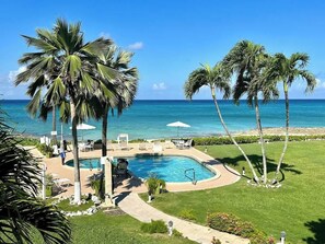 A pool area with lounge chairs and umbrellas, surrounded by palm trees and a view of the ocean.