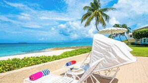 A beachside lounge with white lounge chairs, a small table, and a covered umbrella. - Oceanfront 2 BR & Pool - Cocoplum #8 on 7 Mile (George Town)