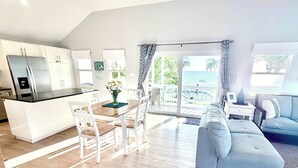 A modern kitchen with white cabinets, a black countertop, and a dining area with a wooden table and chairs. There is a sofa and a small table with a lamp in the living area. - Oceanfront 2 BR & Pool - Cocoplum #8 on 7 Mile (George Town)