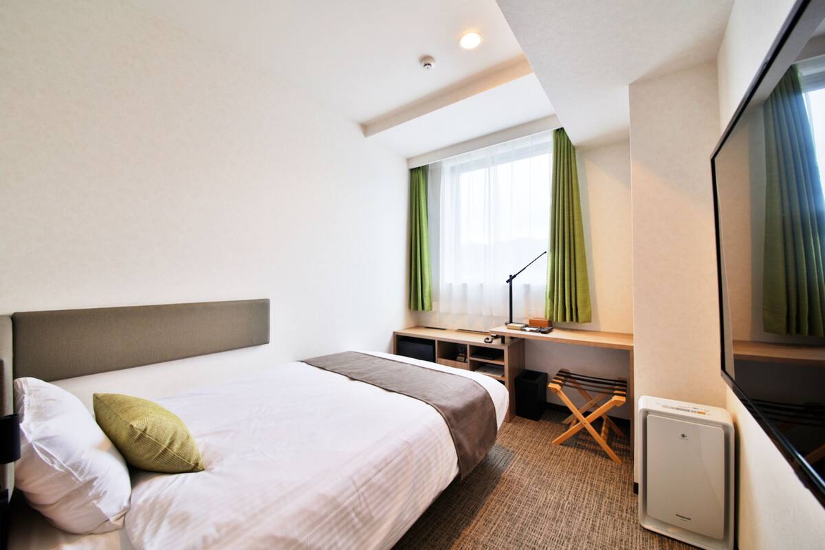 A hotel room with a bed, a desk, a chair, a window with curtains, and a flat-screen TV.