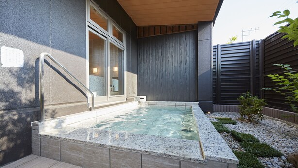 A modern outdoor hot tub area with a stone border, a wooden deck, and a small garden with gravel and plants.