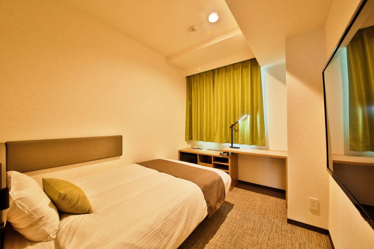 A hotel room with a bed, a desk, a chair, a lamp, a mirror, and a window with curtains.
