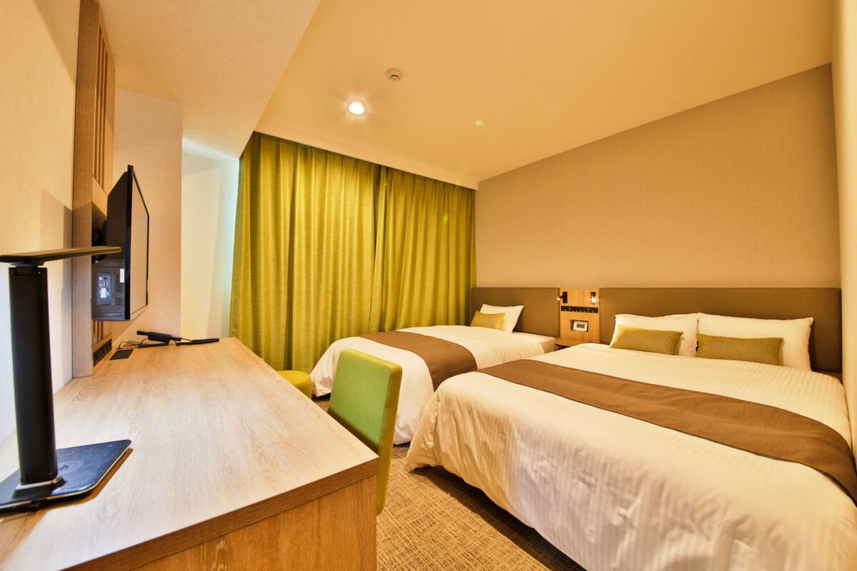 A hotel room with two beds, a desk, a chair, a television, and a green curtain.