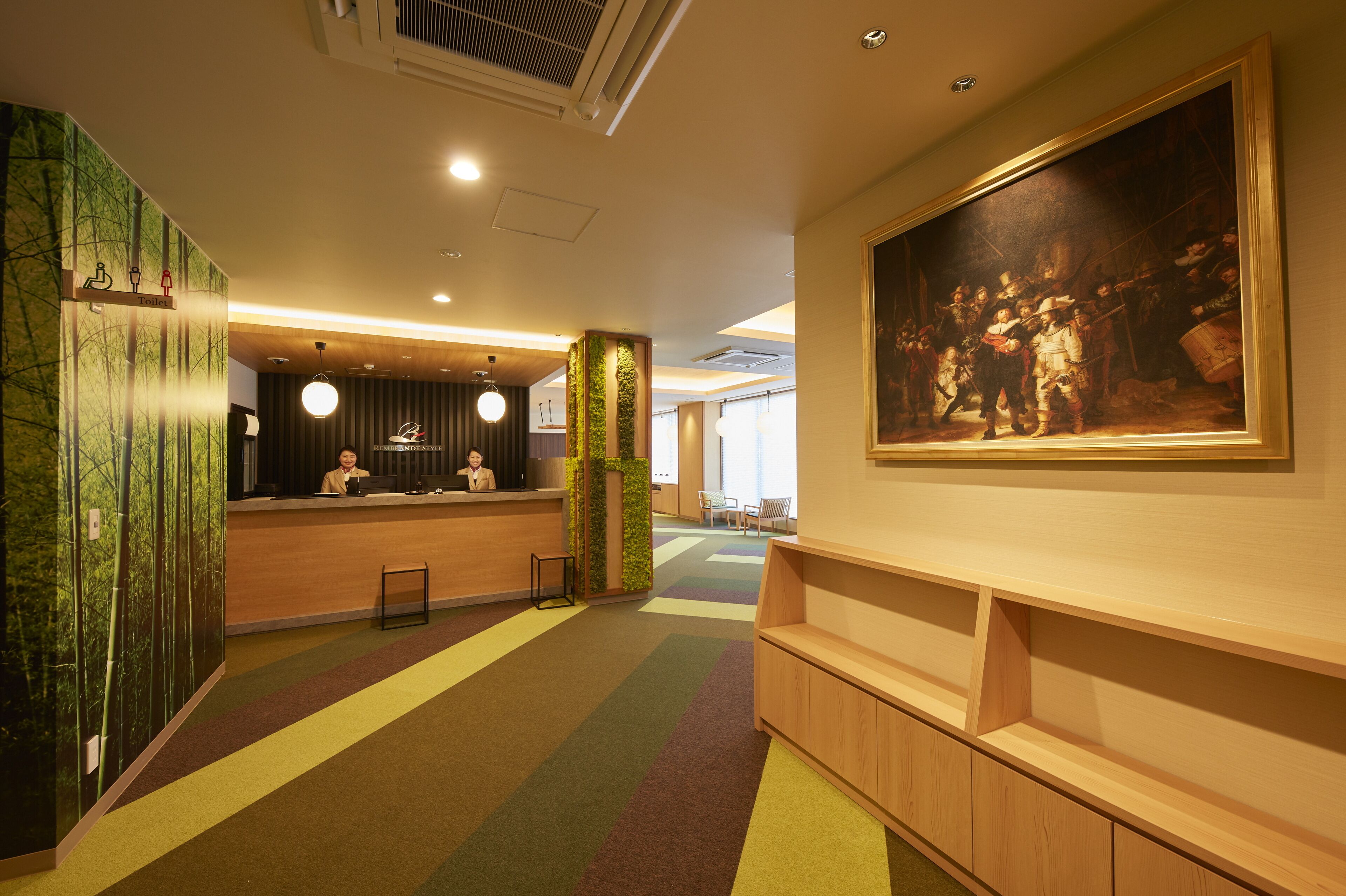 A hotel lobby with a reception desk, a large painting on the wall, and a colorful carpet.