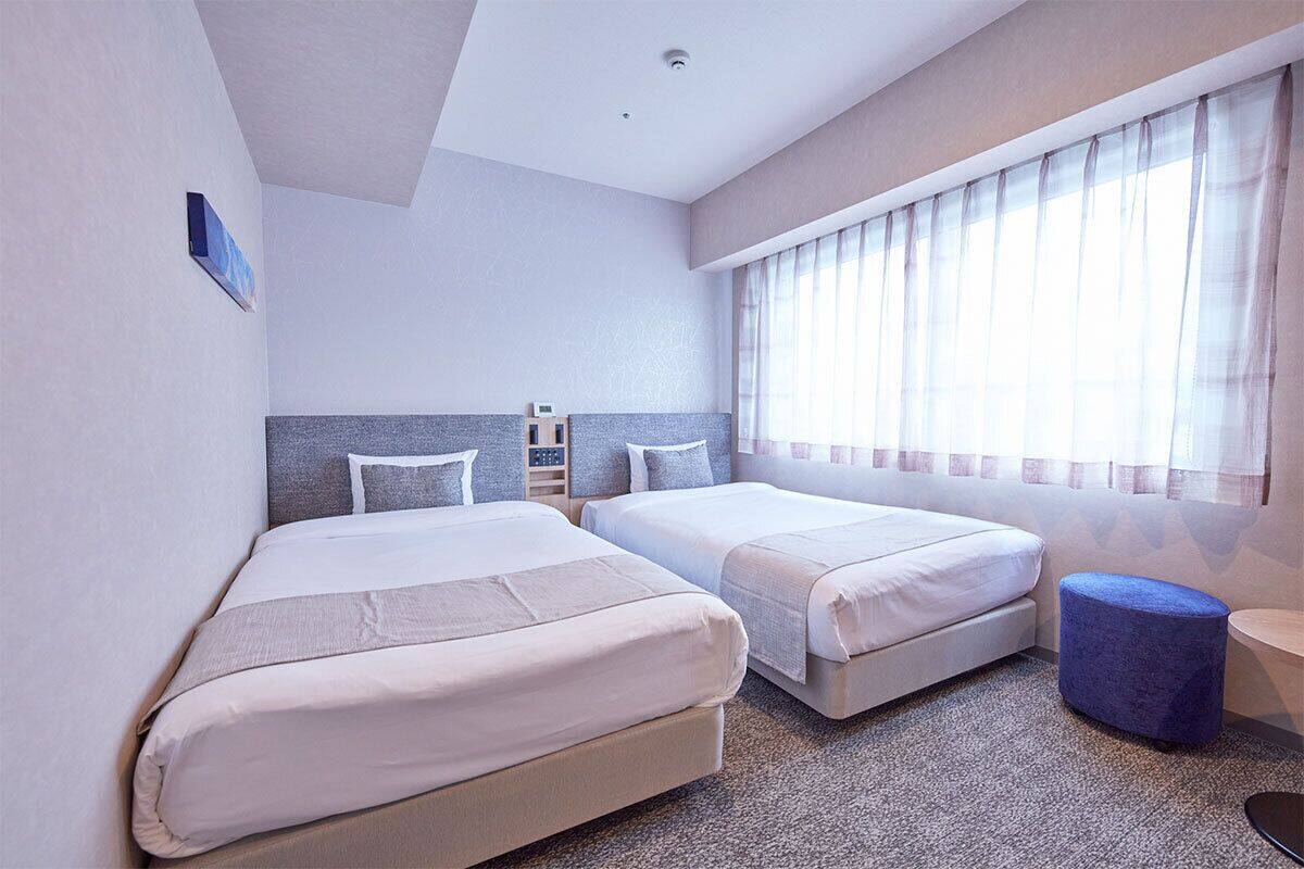 A hotel room with two beds, a small table, and a window with sheer curtains.