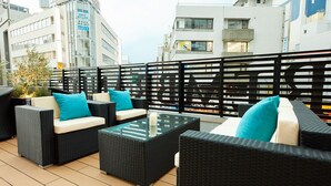 An outdoor seating area with wicker furniture and cushions, overlooking a cityscape with buildings and a parking sign. - Rembrandt Style Honatsugi (Atsugi)