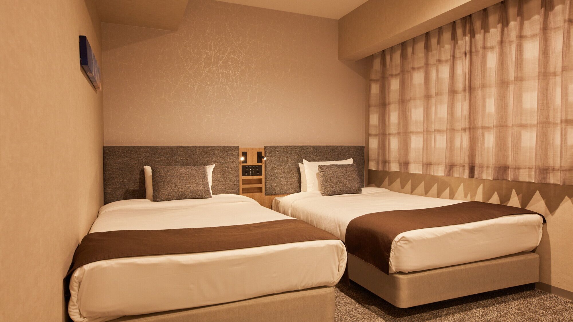 A hotel room with two beds, a headboard with a control panel, a window with curtains, and a wall-mounted artwork.