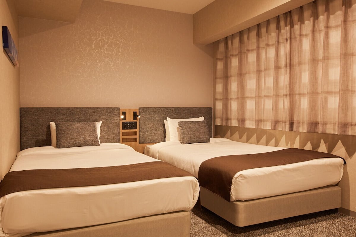 A hotel room with two beds, a headboard with a control panel, a window with curtains, and a wall-mounted artwork.