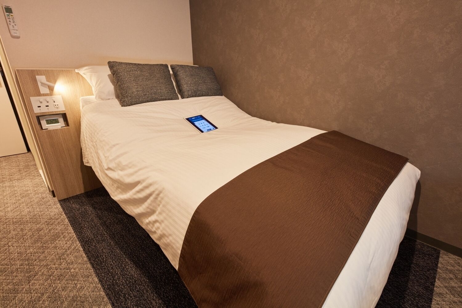 A hotel room with a bed, a bedside table with a clock, and a wall-mounted control panel.