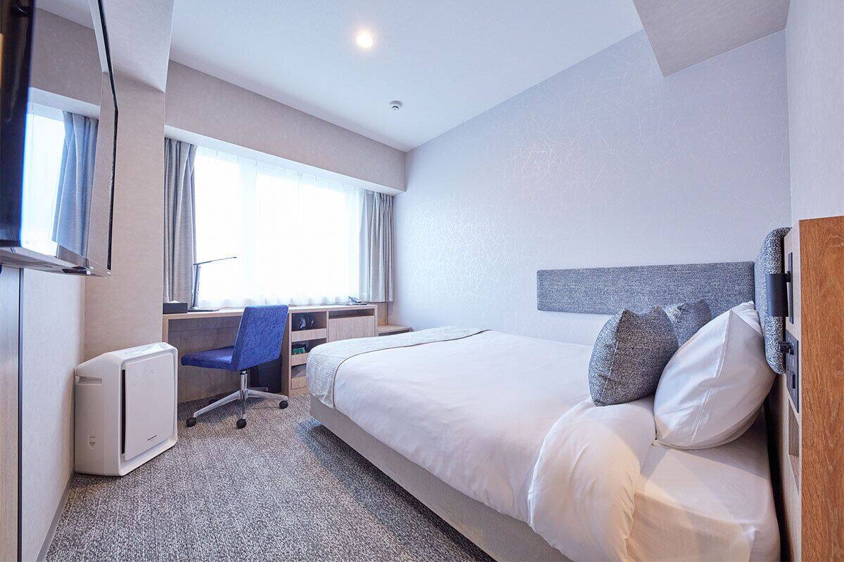A hotel room with a bed, a desk with a chair, a flat-screen TV, and a window with curtains.