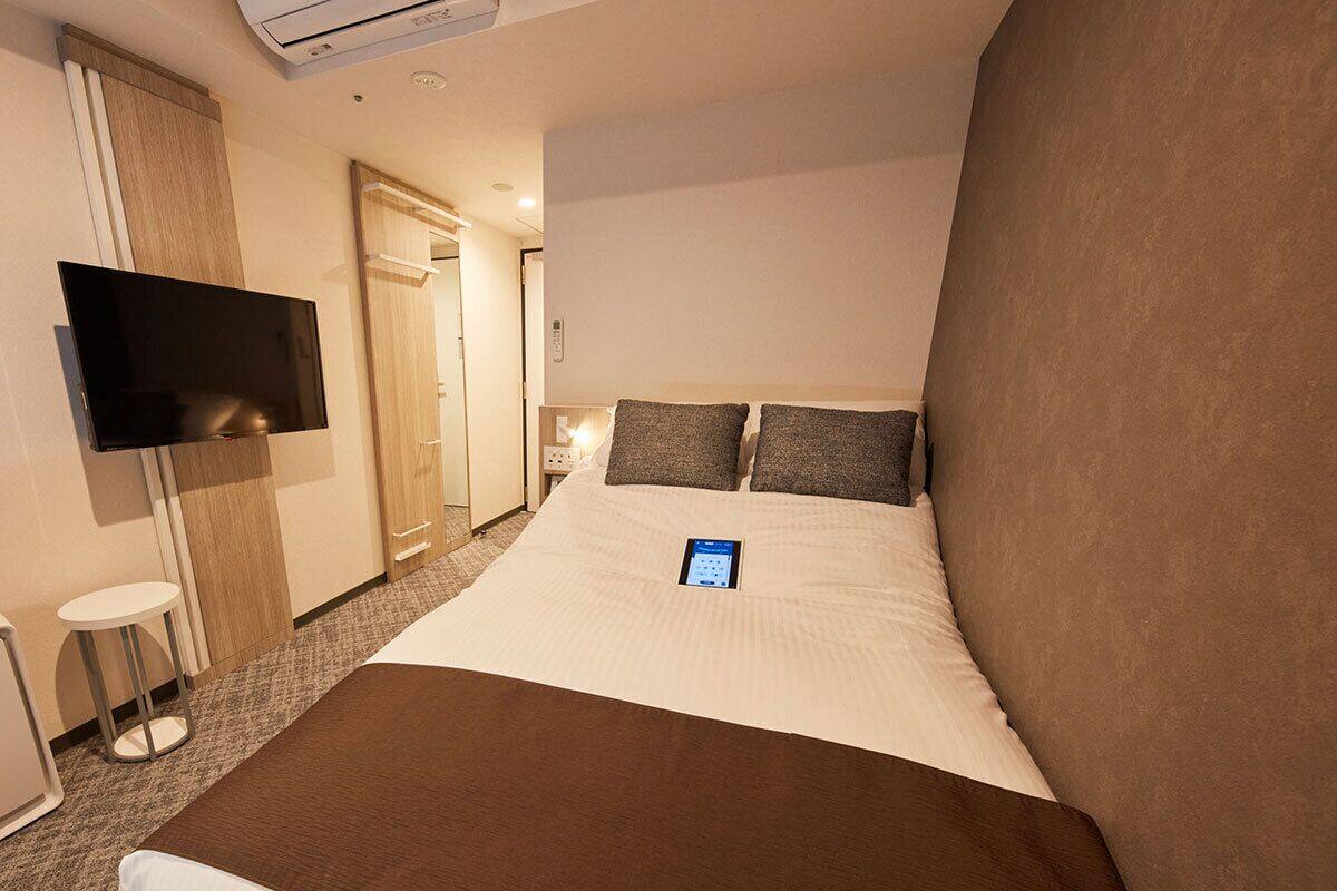 A hotel room with a bed, a flat-screen TV mounted on the wall, a bedside table, and a small stool.