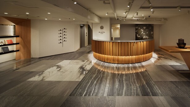 Zona de recepción de un hotel con un mostrador de madera, un panel informativo en la pared y suelo de baldosas.