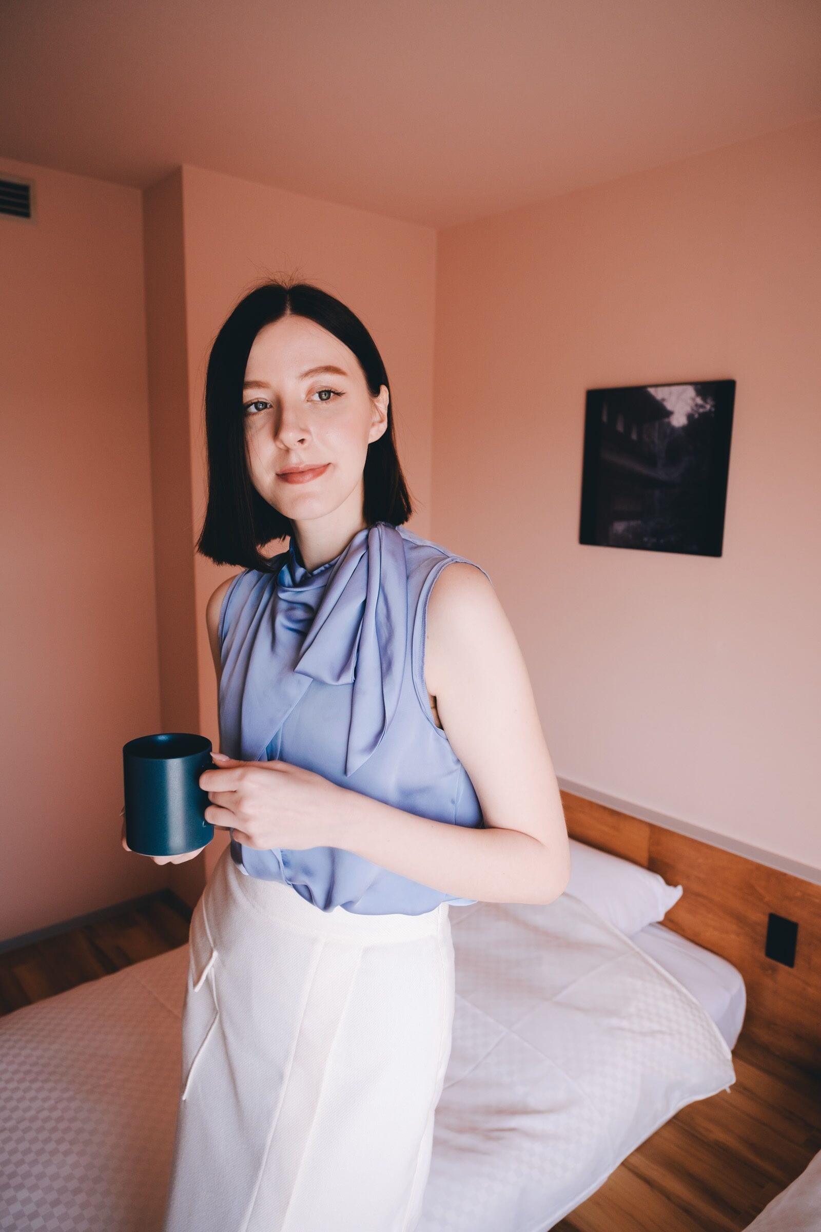 Una persona de pie en una habitación con una cama, sosteniendo una taza.