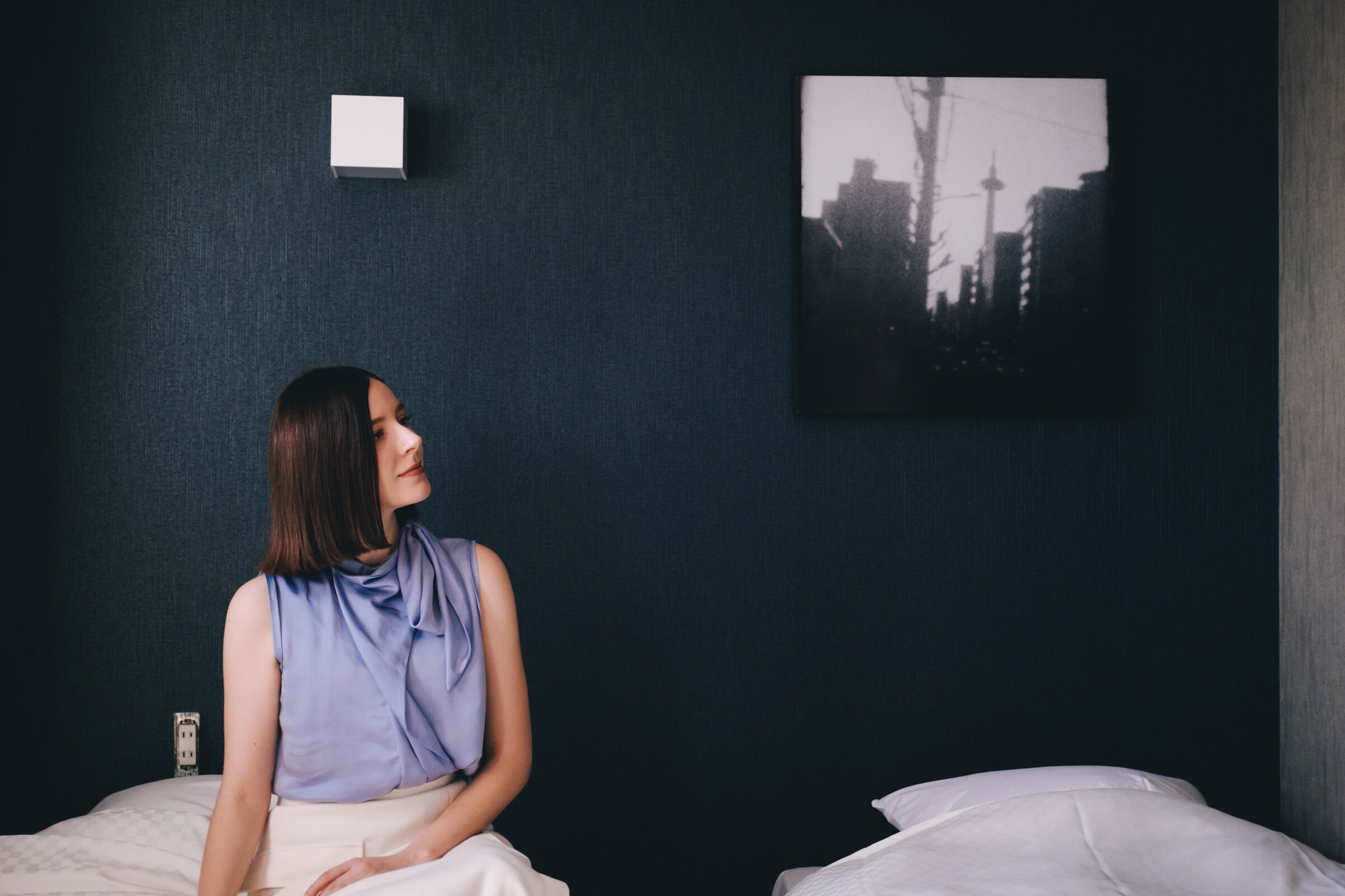 A person sitting on a bed in a room with a dark wall and a cityscape painting.