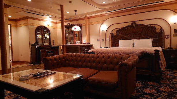 A hotel room with a large bed, a brown tufted sofa, a wooden nightstand, and a mirror. - Hotel Zips - Adults Only (Kawaguchi)