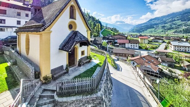 Une petite église dotée d’un clocher, entourée d’un village avec des maisons et surplombée par une montagne.