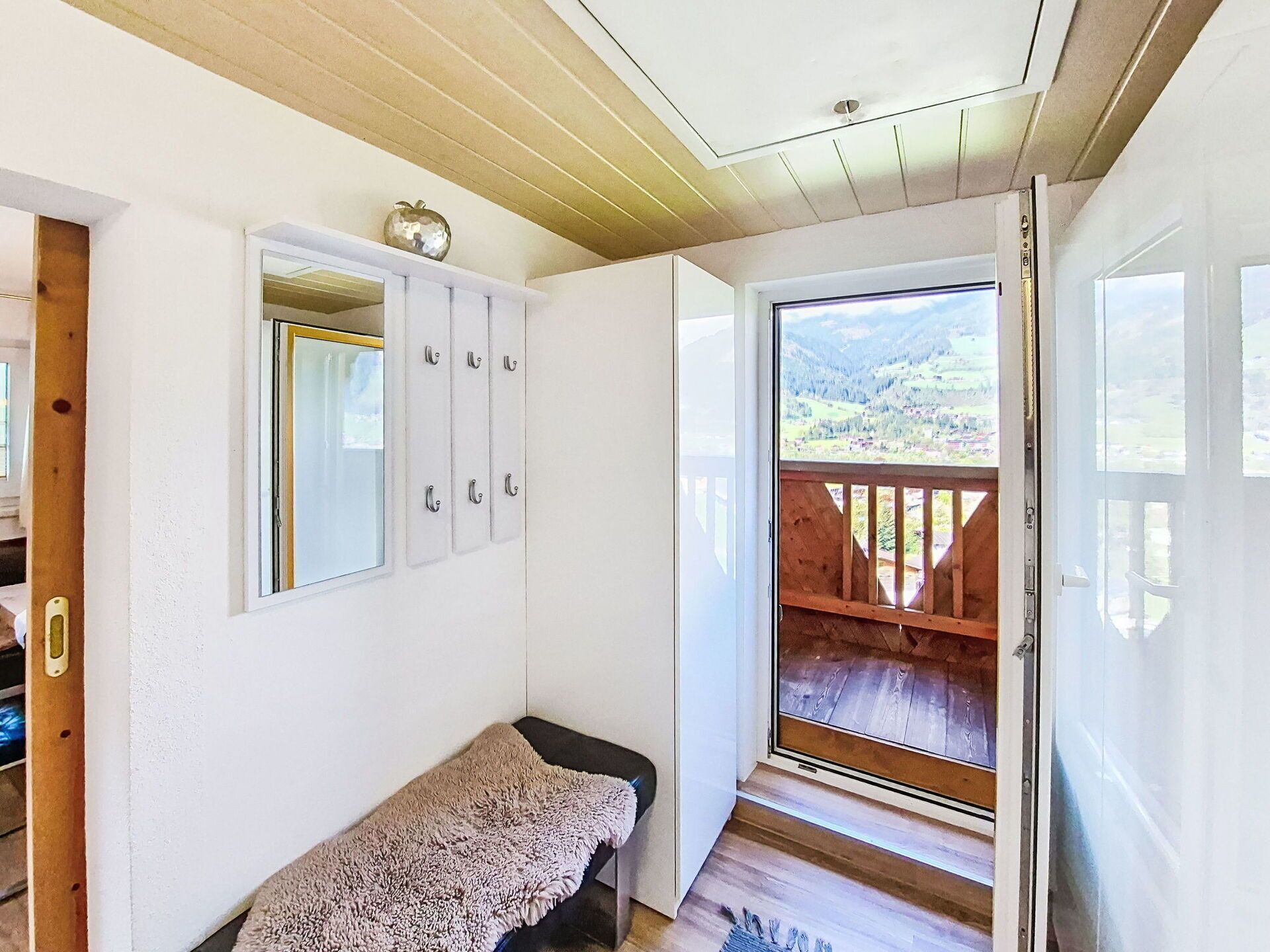 Quarto com teto de madeira, um banco, parede branca com espelho e armários, e uma porta que dá para uma varanda com vista para uma paisagem montanhosa.