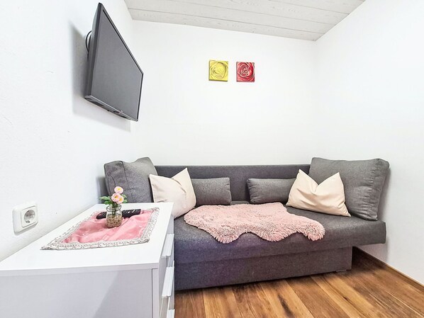 Un salon moderne avec un canapé gris, une table d’appoint blanche, un plaid rose et un téléviseur à écran plat fixé au mur.