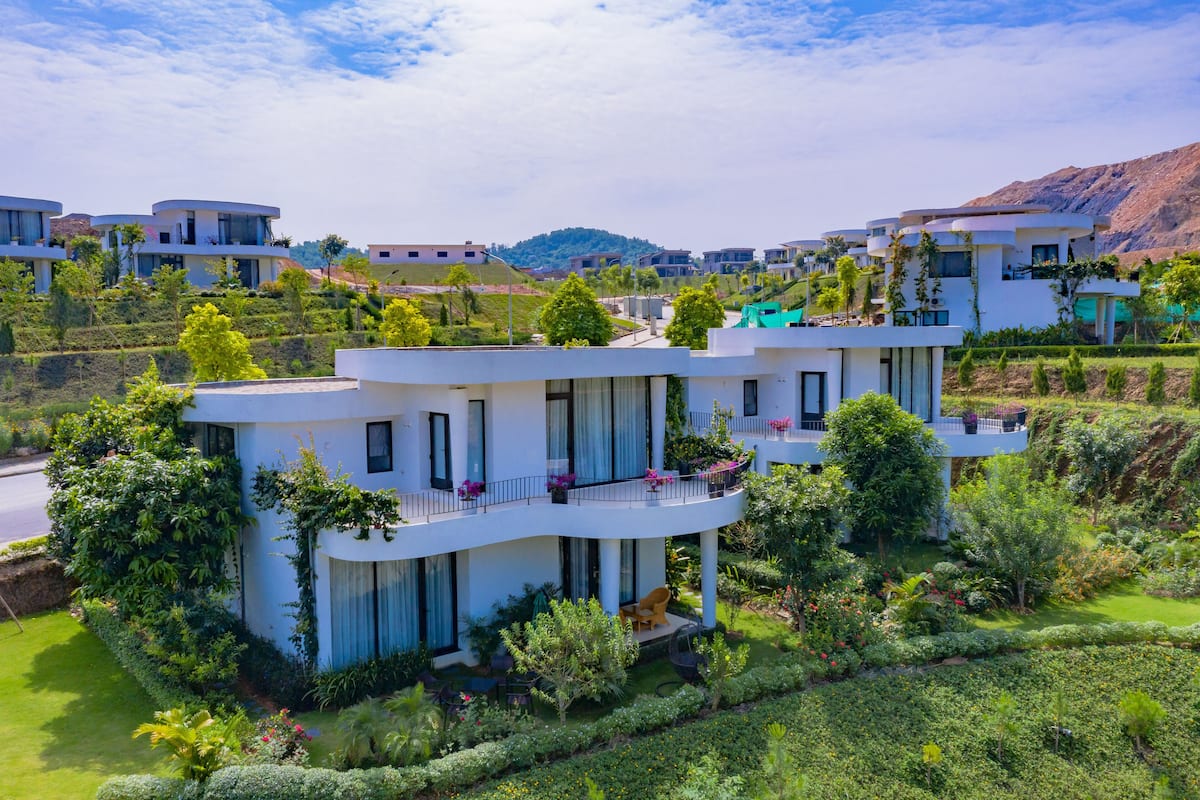 A modern multi-story residential building with balconies, surrounded by lush greenery and a hilly landscape.