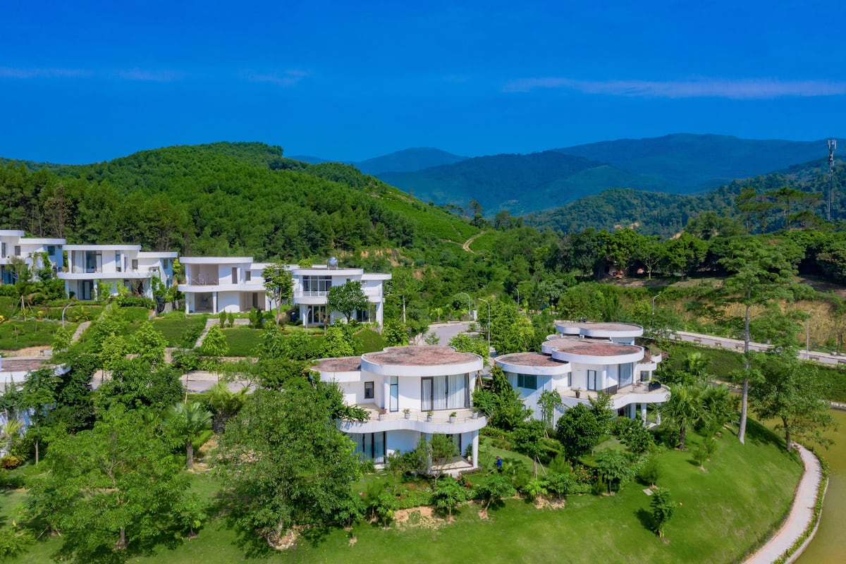 A modern resort with multiple white buildings, lush greenery, and a mountainous backdrop.