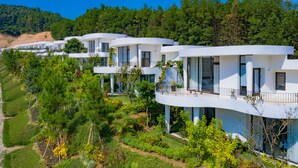 Una hilera de villas modernas y blancas de dos pisos, con balcones, ubicadas en un paisaje exuberante y verde con una ladera boscosa al fondo.