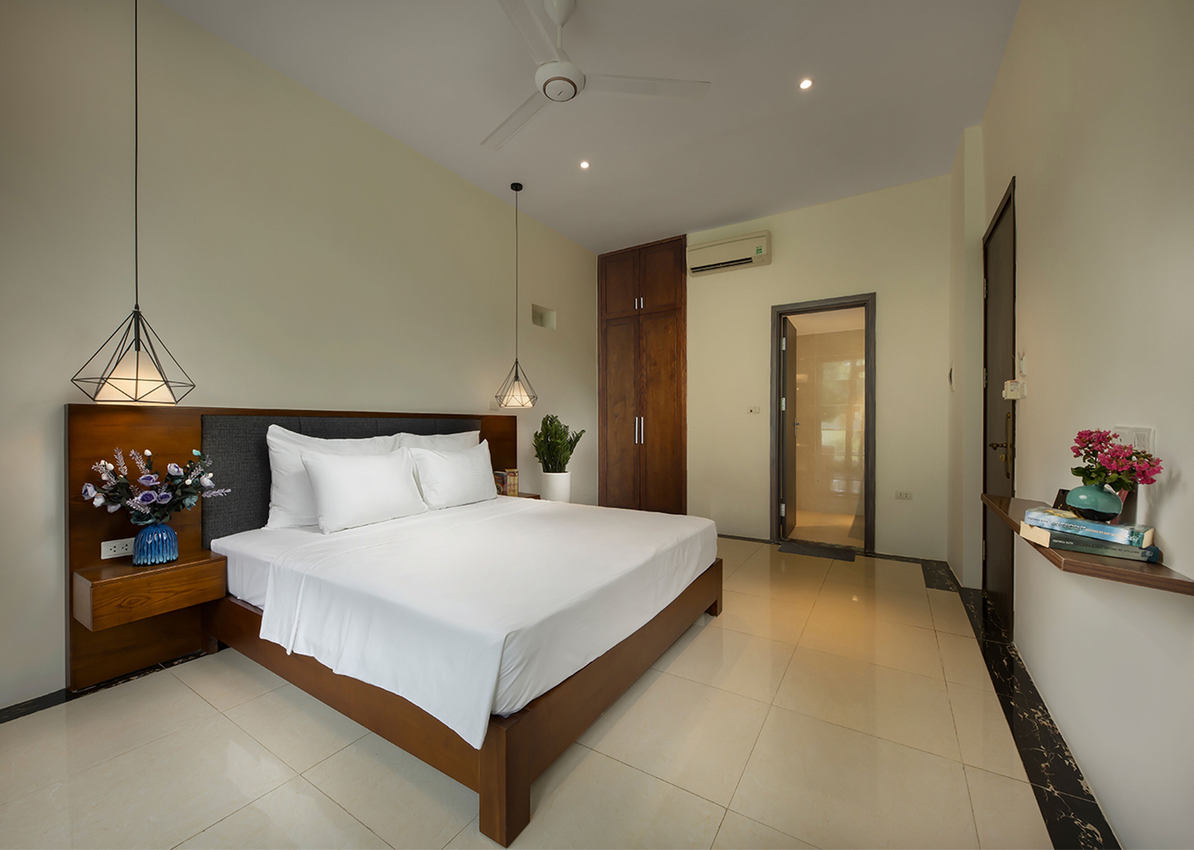 A modern hotel bedroom with a large bed, wooden headboard, bedside tables with lamps, an air conditioning unit, and a door leading to another room.