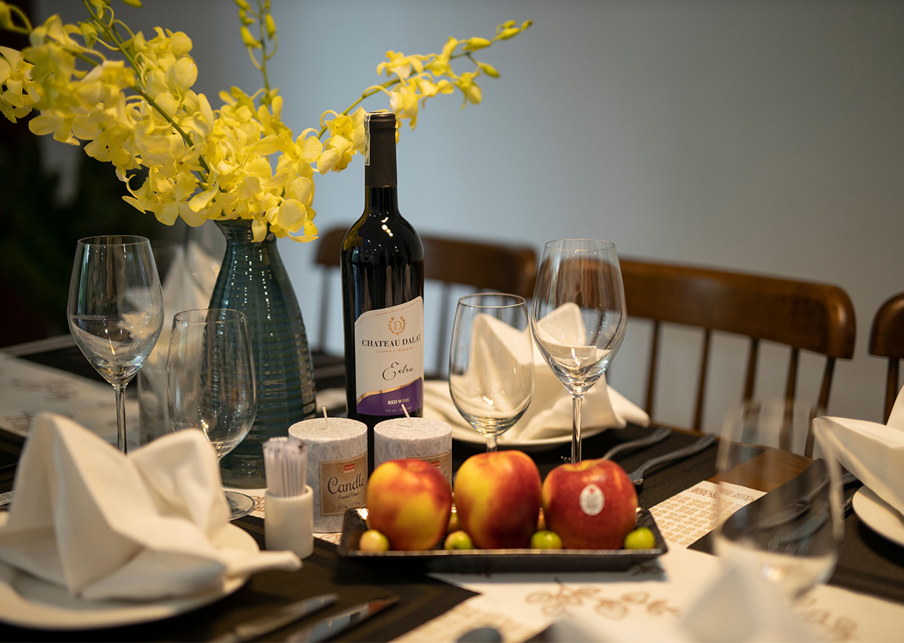 A dining table with a bottle of Chateau Dalat wine, three glasses, a vase with yellow flowers, and a plate with apples.