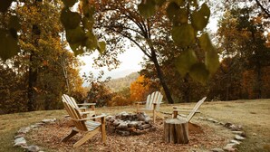 A circular seating arrangement with wooden chairs around a stone fire pit in a grassy outdoor area with trees in the background. - Mt. Sherman Cabin - Ozark Mountains (Jasper)