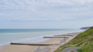 Une plage côtière avec une jetée en bois qui s’avance dans la mer, une rive sablonneuse et une colline herbeuse.