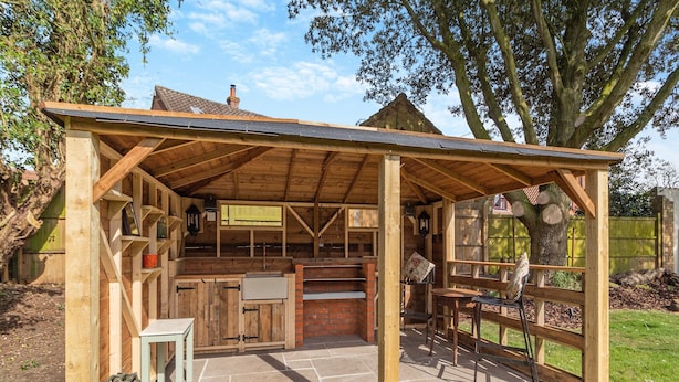Une structure de cuisine extérieure en bois, dotée d’un comptoir en briques, d’un évier et d’un espace repas avec des chaises.