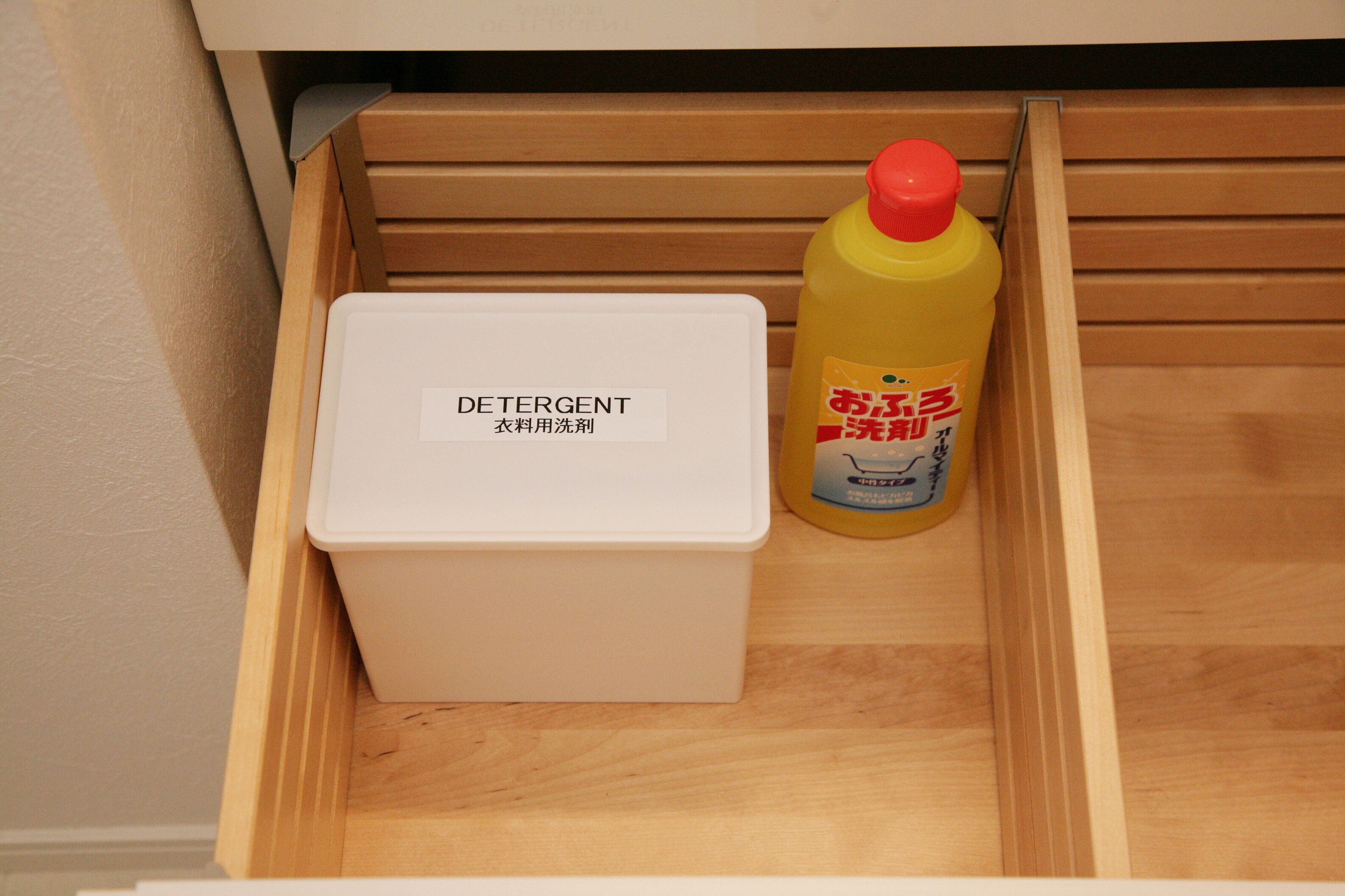 A wooden drawer containing a white detergent container and a yellow bottle with a red cap.