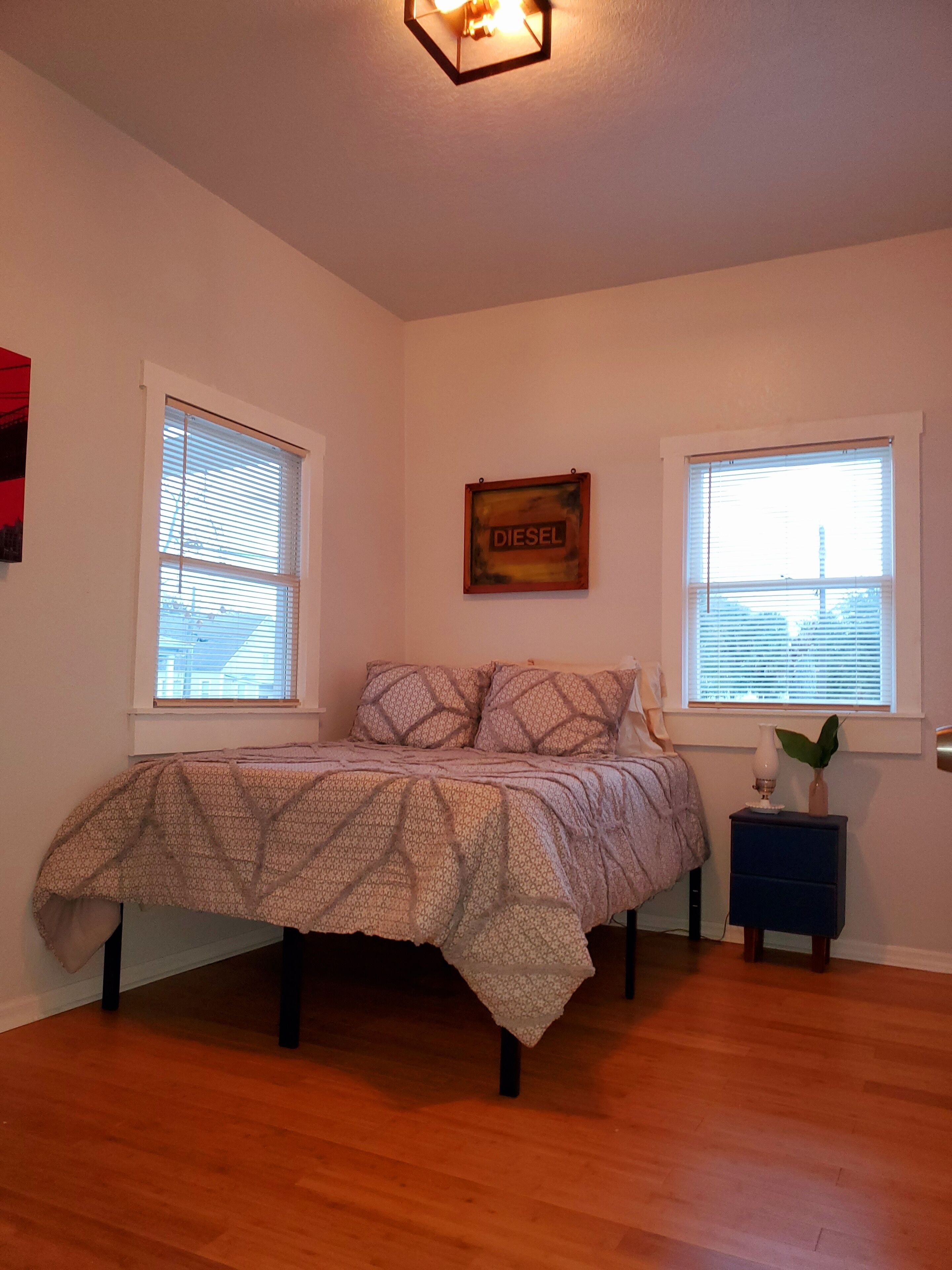 A bedroom with a bed, two windows, a wall-mounted sign, and a small table with a plant.