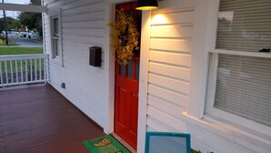Een wit huis met een rode deur, een veranda met een groene deurmat en een blauwe stoel.