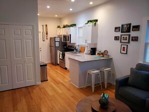 Una cocina moderna con armarios blancos, microondas integrado y una isla central. Hay dos taburetes blancos y un sofá gris en la sala de estar.