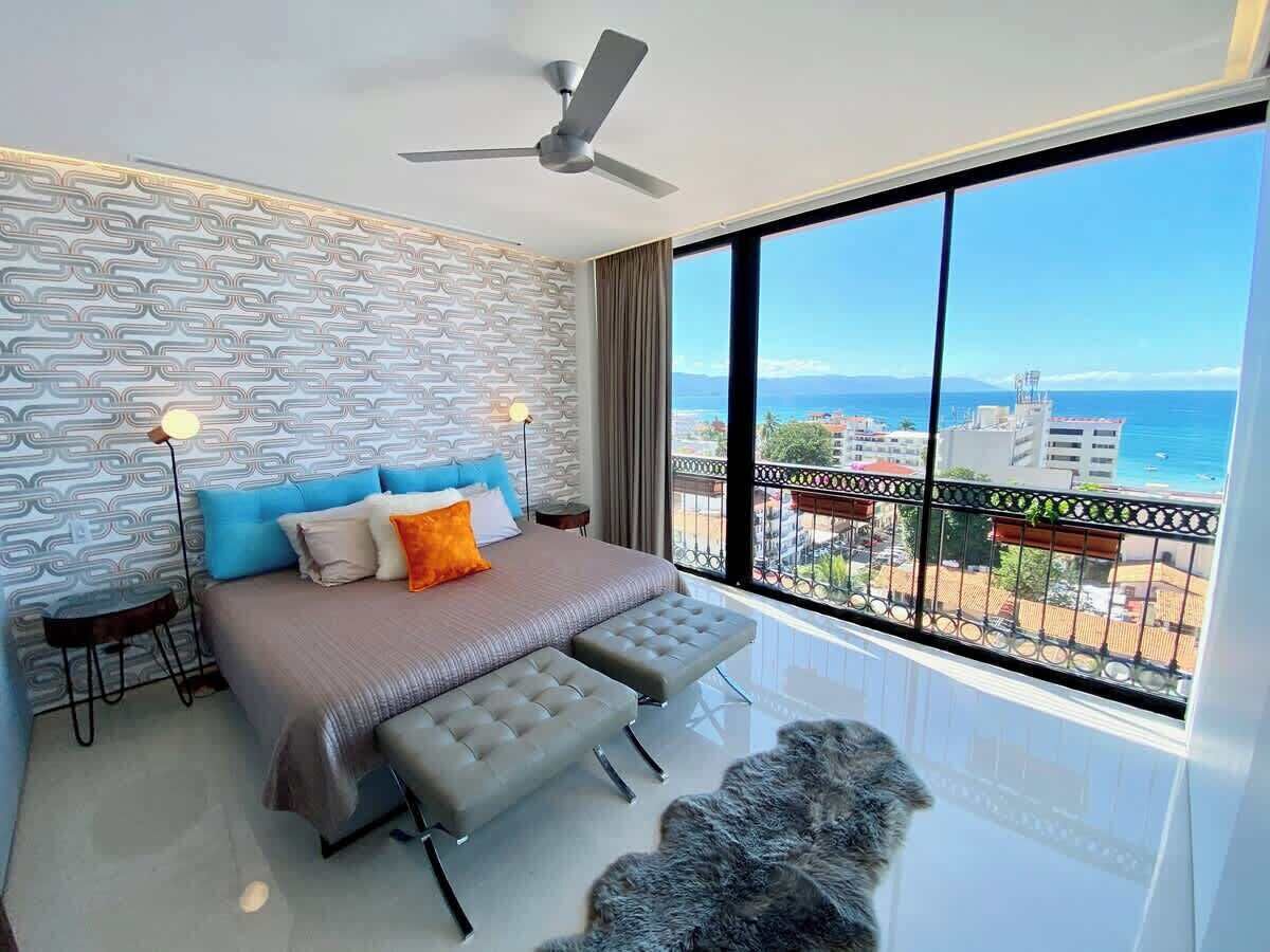 A modern bedroom with a bed, two side tables, a floor lamp, a ceiling fan, and a large window with a view of the city and sea.