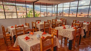 A restaurant with multiple tables and chairs, each set with a tablecloth and orange napkins. Large windows offer a view of a cityscape and mountains. -  HOTEL MOSOQ KILLA (Cusco)