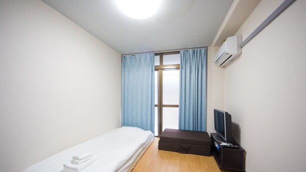 Una habitación pequeña y limpia con una cama, una maleta, un televisor y un aire acondicionado.